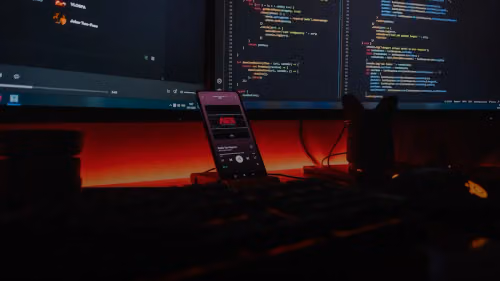 A smartphone displaying music on a desk with computer monitors showing code