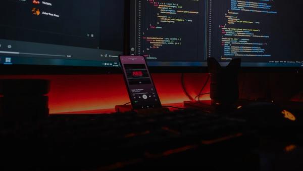 A smartphone displaying music on a desk with computer monitors showing code