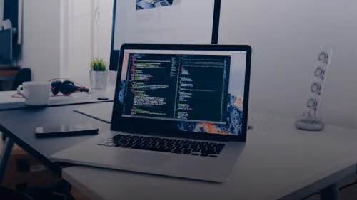 A MacBook with lines of code on its screen on a busy desk